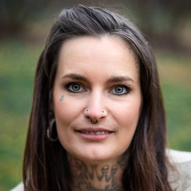 Woman with tattoos and piercings wearing a beige sweater and pearl necklace against a blurred natural background